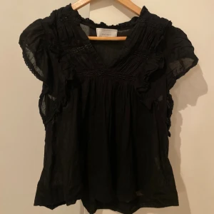 Svart blus med volang från Neo Noir - Svart blus från Neo Noir med v-ringning, volangdetaljer och rynkade partier upptill. Blusen är ärmlös med lätt transparent tyg och har en luftig, loose passform. ❤️❤️