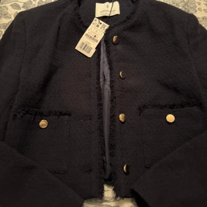 Svart kavajjacka från Stradivarius - Stilren svart kavajjacka från Stradivarius i storlek L. Jackan har guldiga knappar, två fickor framtill och råa kanter för en trendig look. Fodrad insida i glansigt tyg och klassisk rak passform. Perfekt för dig som vill ha en chic och modern stil.