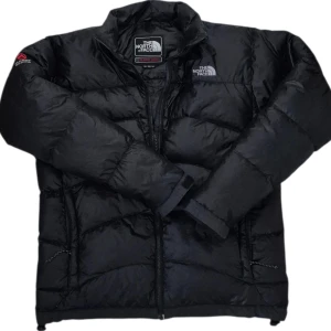 Svart dunjacka från The North Face MEN LARGE - Svart dunjacka från The North Face med klassisk puffer-design. Jackan har hög krage, dragkedja framtill och två sidofickor. Logga på bröstet och ärmen. Perfekt för kalla dagar och riktigt snygg till streetwear-stilen.