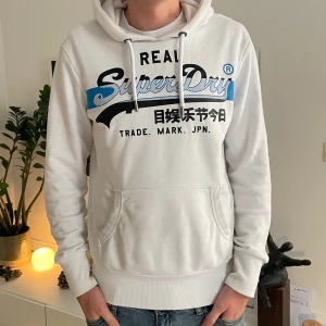 Vit Superdry hoodie med tryck - Vit hoodie från Superdry med svart och blått tryck framtill samt japanska tecken. Klassisk känguruficka, snörning i huvan och ribbade muddar. Perfekt för dig som gillar streetwear och vill ha en snygg och bekväm tröja.