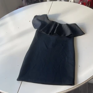 Svart one shoulder klänning - Snygg svart kortklänning med one shoulder och stor volangdetalj över axeln. Klänningen har en tight passform och är perfekt för dig som vill sticka ut. Enkel men ändå trendig design som passar till många tillfällen.