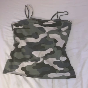 Camo linne med smala axelband - Säljer ett camouflagemönstrat linne i gröna, beige och vita toner. Toppen har smala, justerbara axelband och är figurnära i modellen. Perfekt för dig som gillar streetstyle och vill sticka ut med ett edgy mönster.