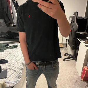 Svart t-shirt från Polo Ralph Lauren - Svart t-shirt från Polo Ralph Lauren med klassisk röd broderad logga på bröstet. T-shirten har rund halsringning och korta ärmar. Tillverkad i mjuk bomull för en skön och avslappnad passform.