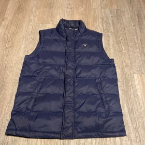 Marinblå dunväst från Gant - Snygg marinblå dunväst från Gant med quiltad design och ståkrage. Västen har två snedställda fickor framtill och dragkedja. Insidan är fodrad med blåvitrandigt tyg för en fräsch look. Perfekt för lager-på-lager under kyliga dagar.