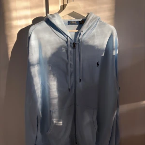 Ljusblå hoodie från Polo Ralph Lauren - Säljer en ljusblå hoodie från Polo Ralph Lauren med dragkedja och snörning i huvan. Klassisk logga broderad på bröstet och två fickor framtill. Perfekt för dig som gillar stilrena och bekväma plagg.