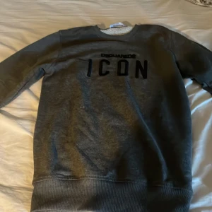 Grå sweatshirt från Dsquared2 ICON - Säljer en grå sweatshirt från Dsquared2 med svart 'ICON'-tryck på bröstet. Tröjan har rund halsringning, långa ärmar och breda ribbade muddar vid ärmslut och nederkant. Perfekt för dig som gillar streetwear och vill sticka ut med ett ikoniskt plagg.