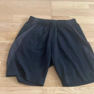 Svarta träningsshorts med grå detalj - Svarta träningsshorts med grå paneler på sidorna och elastisk midja. Liten logga nere på benet. Lätta och luftiga, perfekta för gymmet eller löprundan. Klassisk loose fit och sportig look.