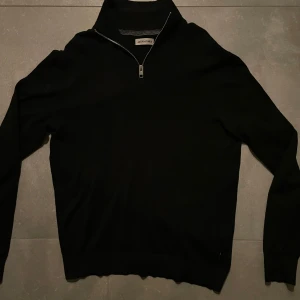 Svart half zip-tröja Jack & Jones - Svart långärmad tröja från Jack & Jones med hög krage och half zip-dragkedja. Enkel och stilren design, perfekt för lager-på-lager. Mjuk och bekväm känsla, passar till många olika outfits. Storlek s, litet hål längst ner till höger men inget man märker