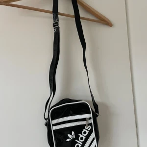 Svart axelväska från Adidas - Cool svart axelväska från Adidas med vit logga och detaljer. Väskan har justerbar axelrem, dragkedja och är tillverkad i glansigt syntetmaterial. Perfekt för att bära dina viktigaste grejer och ge din outfit en sportig touch.