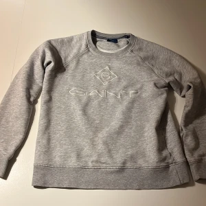 Grå sweatshirt från GANT - Säljer en stilren grå sweatshirt från GANT med broderad logga framtill. Tröjan har rund halsringning och långa ärmar. Perfekt för dig som gillar klassisk och enkel stil. Materialet är mjukt och skönt, passar till många olika outfits.