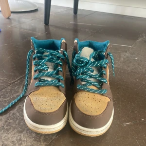 Nike Air Jordan 1 Mid brun/blå - Säljer ett par Nike Air Jordan 1 Mid med brun mocka och skinn, beige detaljer och turkosa snören. Skorna har en klassisk Jordan-silhuett med perforerad tå och blått innerfoder. Perfekta för dig som vill sticka ut med en unik färgkombo.