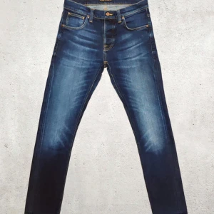 Nudie Jeans 31/32 - Säljer ett par blå skinny jeans från Nudie Jeans med klassisk femficksdesign och snygga slitningar framtill och baktill. Jeansen har orangea sömmar och är tillverkade i stretchig denim för en tight passform. Perfekta för dig som gillar en modern och smal siluett.