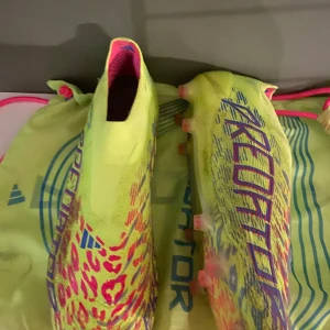 Adidas Predator fotbollsskor neon elit - Säljer ett par Adidas Predator fotbollsskor i neon-gult med lila och rosa detaljer samt leopardmönster. Skorna har en strumpliknande passform utan snörning och är tillverkade i syntetmaterial. Perfekta för dig som vill sticka ut på planen!
