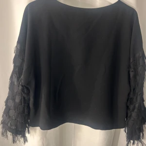 Svart blus med fransiga ärmar H&M - Svart blus från H&M i storlek 36 med vida, fransiga ärmar som har ett coolt mönster av cirklar och fransdetaljer. Toppen har en rak, croppad passform och rund halsringning. Perfekt för dig som vill ha något unikt och trendigt i garderoben.