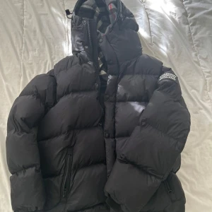 Svart pufferjacka från Burberry - Säljer en svart pufferjacka från Burberry med klassiskt rutigt foder i huvan. Jackan har dragkedja, knappar och två fickor med dragkedja framtill. Perfekt för kalla dagar och riktigt snygg med sin puffiga look och ikoniska Burberry-detaljer.