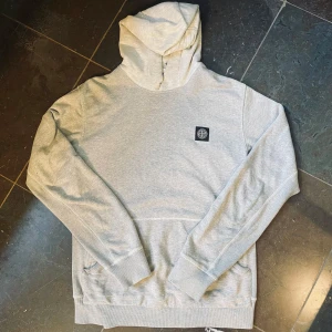 Ljusgrå hoodie från Stone Island - Säljer en stilren ljusgrå hoodie från Stone Island med klassisk logga på bröstet. Tröjan har huva, känguruficka och ribbade muddar.                              📏 Storlek: L (men sitter mer som en M) ✨ Skick: Använd men i bra skick, inga fläckar eller hål. 🧵 Material: 100% bomull – bekväm och slitstark.       ✅ Pris kan diskuteras