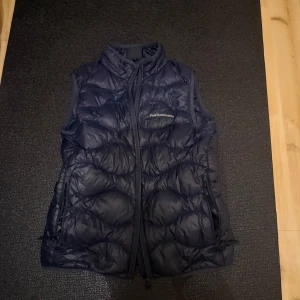 Mörkblå dunväst Peak Performance - Snygg mörkblå dunväst från Peak Performance med quiltad design och hög krage. Västen har dragkedja framtill och två sidofickor med dragkedja. Perfekt att slänga över en hoodie när det är kyligt ute.