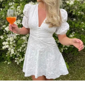 Adoore klänning - Superfin vit kortklänning med puffiga kortärmar och v-ringad hals. Klänningen har ett subtilt blommigt mönster och markerad midja med knappar framtill. Perfekt för studenten!!