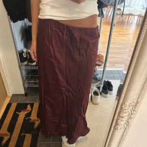 Vinröd långkjol i satin - Säljer en vinröd långkjol med mjukt satintyg och lätt glansig finish. Kjolen har en enkel, rak siluett och sitter snyggt i midjan. Perfekt för dig som gillar stilrena plagg med lite extra lyxig känsla.