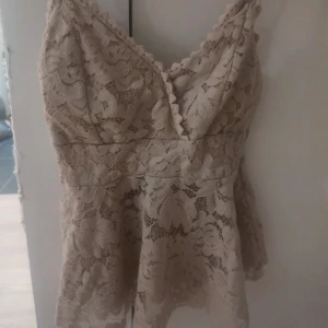 Beige spetslinne med blommönster - Säljer ett superfint beige linne med v-ringning och breda axelband. Toppen är täckt av vacker spets med blommönster och har en lätt utsvängd passform. Perfekt för dig som gillar romantisk och feminin stil.