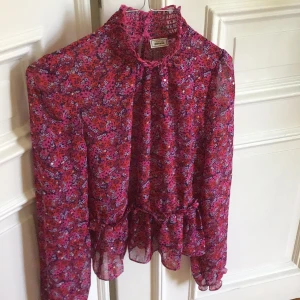 Blommig volangblus från Pimkie - Supersöt blommig blus från Pimkie i rosa, lila och rött med volangdetaljer och hög krage. Blusen är transparent, har långa ärmar och knappar bak i nacken. Perfekt för dig som gillar romantiska och färgglada plagg.