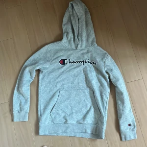 Grå hoodie från Champion - Snygg grå hoodie från Champion med broderad logga på bröstet och liten logga på ärmen. Klassisk känguruficka framtill, mjukt material och huva. Perfekt för chill dagar eller när du vill ha en sportig look.