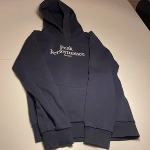Mörkblå hoodie från Peak Performance - Säljer en mörkblå hoodie från Peak Performance med broderad logga på bröstet. Tröjan har huva, magficka och ribbade muddar vid ärmslut och nederkant. Perfekt för dig som gillar en clean och sportig stil.