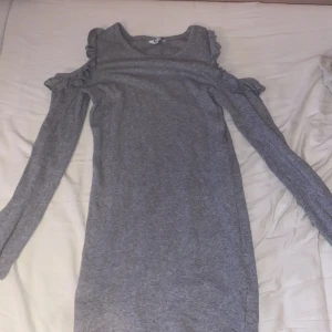 Grå ribbad klänning med volang - Grå ribbad klänning med långa ärmar och snygga volangdetaljer vid axlarna. Modellen har cold shoulder-design som ger en cool touch. Perfekt för dig som gillar en enkel men ändå trendig stil.