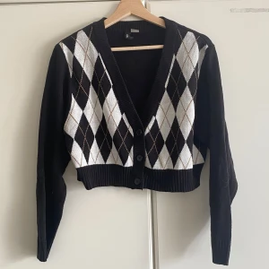 Svart och vit rutig kofta med knappar - Snygg croppad kofta från h&m i svart med klassiskt rutigt argylemönster i vitt och beige framtill. Djup v-ringning, ribbade muddar och tre knappar framtill. Perfekt för lager-på-lager och ger en preppy vibe.