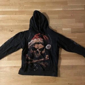 Svart hoodie med dödskalle och tomteluva - Svart hoodie med stor dödskalle och korslagda ben på framsidan, båda med tomteluva i rött och vitt. Baksidan har en dödskalle med tomteluva och coola ringar. Ärmarna har tryckta flames. Huva med snörning och känguruficka framtill. Nypris 699kr, endast använd 2 gånger 