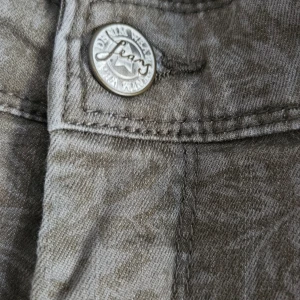 Olivgrön blommig jeans  - Snygga grå jeans från kappahl med subtilt blommönster över hela byxan. Modellen har klassisk femficksdesign och raka ben. Jeansen är tillverkade i bomull och har en cool, lite tvättad look som sticker ut. Storlek 42. Normal hög midja