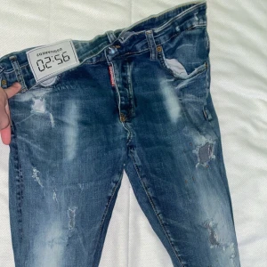 Blå slitna jeans från Dsquared2 - Säljer ett par blå jeans från Dsquared2 med slitningar och trasiga detaljer på flera ställen. Jeansen har klassisk femficksmodell, knappgylf och en röd Dsquared2-tag vid gylfen. Snyggt tvättade och med en modern, smal passform.