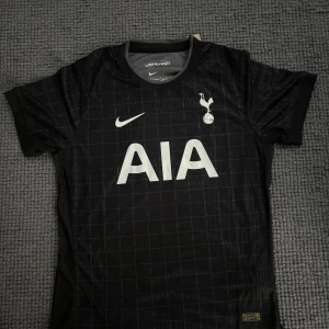 Tottenham svart matchtröja Nike L - Snygg svart Tottenham Hotspur fotbollströja från Nike med diskret rutmönster och stora vita tryck på bröstet. Tillverkad i Dri-FIT ADV-material som andas. Klassisk rund hals och korta ärmar, perfekt för dig som älskar Spurs.