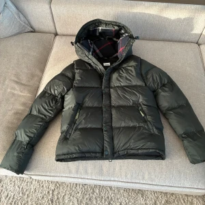  pufferjacka från Burberry - Säljer en pufferjacka från Burberry med klassiskt rutigt foder i huvan. Jackan har dragkedja och knappar framtill, två dragkedjeförsedda fickor och en mysig huva. Perfekt för kalla dagar och riktigt snygg streetstil. Man kan också dra av armarna så den blir till en väst vilket är perfekt nu till hösten och sen till vintern!🍁 Kom med frågor om det är nått ni vill fråga! Pris kan diskuteras!