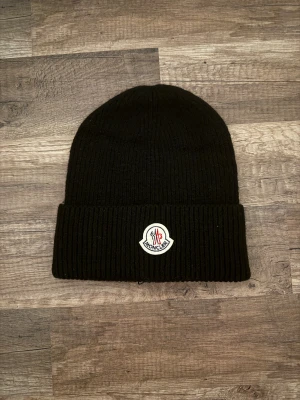 Svart Moncler mössa i ull - Svart ribbstickad mössa från Moncler med uppvikt kant och klassisk Moncler-logga framtill. Mössan är tillverkad i mjuk ull och har en enkel, stilren design som passar perfekt för kalla dagar. Snygg accessoar som lyfter din vinterstil.