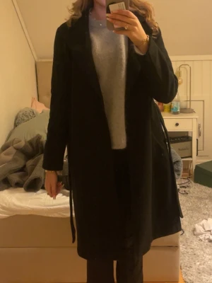 Svart lång kappa med bälte - Säljer en svart, stilren och lång kappa med bälte i midjan. Klassisk rak passform och långärmad design, perfekt för dig som gillar en clean look. Passar dig som vill ha en enkel men snygg jacka till höst och vinter.