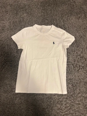 Vit t-shirt från Ralph Lauren - Vit t-shirt från Ralph Lauren med klassisk passform och korta ärmar. Ikoniska lilla Polo-loggan broderad på bröstet. Tillverkad i mjuk bomull som känns skön mot huden. Perfekt basic-plagg med stilren look.