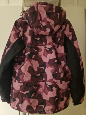 Rosa och vinröd camo vindjacka - Snygg vindjacka från Cross Sportswear i rosa och vinröd camouflage. Jackan har huva med dragsko, svarta paneler på ärmarna och dragkedjor framtill. Perfekt för dig som vill sticka ut med ett coolt mönster och sportig vibe.