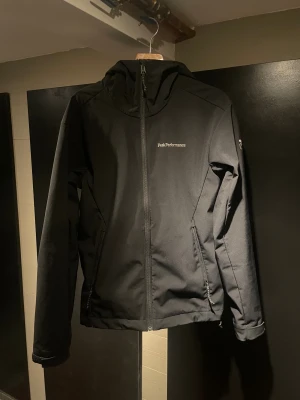Svart vindjacka Peak Performance S - Svart vindjacka från Peak Performance i storlek S. Jackan har huva, hel dragkedja framtill och två sidofickor med dragkedja. Enkel och stilren design med logga på bröstet. Perfekt för blåsiga dagar och passar till många olika outfits.