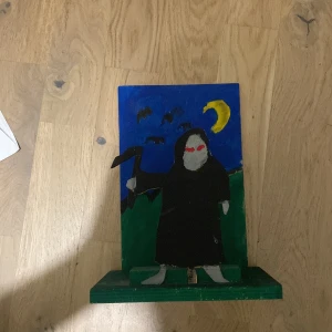 Svart rock med luva, handmålad - Unik handmålad rock i trä, föreställande en figur med svart huva och röda ögon mot en mörkblå bakgrund med måne och fåglar. Rocken har en loose passform och är målad i svart. Perfekt som dekoration eller konstverk.