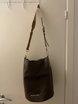 Brun axelväska från Michael Kors - Snygg brun axelväska från Michael Kors med klassiskt MK-mönster och guldfärgade detaljer. Väskan har cylinderformad design, justerbar axelrem i skinn och dragkedja upptill. Insidan är ljusbeige med flera fack och logotyp. Perfekt för dig som gillar stilrena accessoarer.