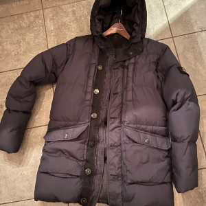 Svart dunjacka från Stone Island - Säljer en svart dunjacka från Stone Island med huva och stora fickor framtill. Jackan har både dragkedja och knappar och en klassisk patch på ärmen. Perfekt för kalla dagar och har en snygg, lite längre passform.