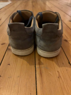 Grå mockasneakers med röd sula - Snygga sneakers i grå mocka med blå snören och vit mellansula. Insidan är beige och sulan har en cool röd undersida. Skorna har rund tå och klassisk snörning, perfekt för dig som gillar stilrena men ändå unika detaljer.