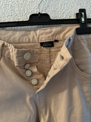 Beige chinos från KappAhl - Snygga beige chinos från KappAhl med hög midja och smal passform. Byxorna har fyra dekorativa knappar i gylfen, klassiska fickor fram och bak samt bälteshällor. Perfekta till en avslappnad men ändå stilren look.
