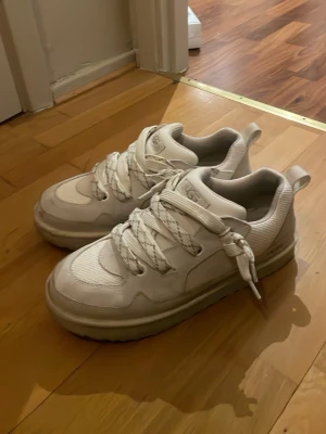 Vita chunky sneakers från UGG - Säljer ett par krämvita UGG lowmell med breda snören och platt sula. Skorna har en mix av skinn och mesh-material, samt UGG-logga på plösen. Snyggt mönstrade snören och grov yttersula ger en cool streetstil. Perfekta för dig som gillar trendiga och bekväma sneakers.