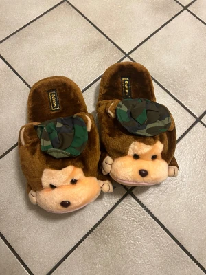 Bruna tofflor med apa och camo-detalj - Mysiga bruna tofflor från Sabina i plysch med gulligt apansikte framtill och en cool kamouflagemönstrad hatt. Perfekta för att chilla hemma och hålla fötterna varma. Sula med halkskydd för extra komfort.