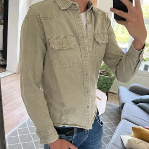 Grön overshirt med knappar - Snygg grön overshirt med klassisk krage och två bröstfickor med lock och knappar. Skjortan har lång ärm och är tillverkad i ett robust bomullsmaterial. Perfekt att bära över en t-shirt för en avslappnad look. Fina detaljer vid sömmarna och knäppning hela vägen.