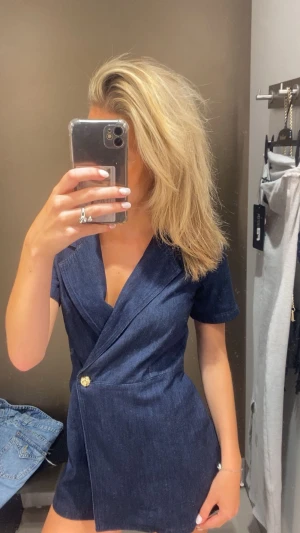 Mörkblå jeansbyxdress med omlott - Snygg mörkblå jeansbyxdress med korta ärmar och omlott-design framtill. Byxdressen har en guldfärgad knapp i midjan och markerad krage. Materialet är jeans och passformen är normal, perfekt för dig som vill ha en cool och modern look.