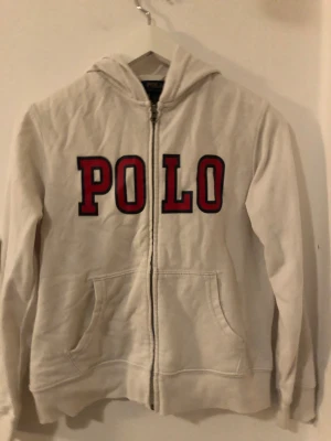 Vit hoodie från Polo Ralph Lauren - Vit hoodie från Polo Ralph Lauren med stor röd POLO-text framtill. Tröjan har dragkedja, huva och två fickor. Tillverkad i mjuk bomull, perfekt för chill dagar. Klassisk och enkel stil som funkar året runt.