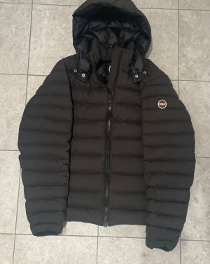 Colmar Down jacket - Svart pufferjacka från Colmar med huva och snygga breda kanaler. Jackan har dragkedja framtill och Colmar-logga på vänster ärm. Perfekt för kalla dagar. Sitter bra på mig som är 185 skulle säga att strl:52 motsvarar en liten medium. Ny-pris: 5500kr.